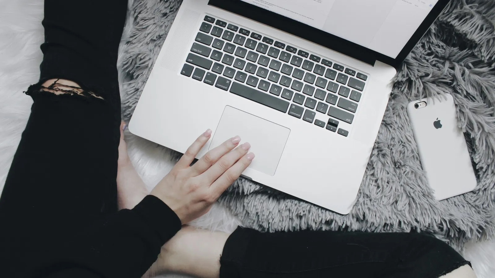 Overhead view of a person in black ripped jeans using a laptop on a grey rug, with a white iPhone beside it